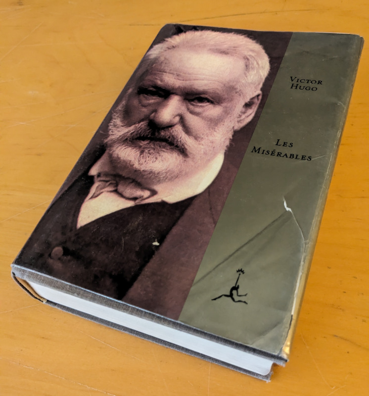 A thick hardcover book, its wraparound cover showing a black and white portrait of Victor Hugo (at the time an old man with white hair and a neatly trimmed beard). The cover is also wrapped in a clear plastic sheet.
