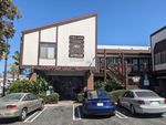 The same building seen from the side. The roof slants up diagonally away from the street. Behind it (to the right, in this view) the building opens into a three-sided courtyard, with wooden stairs up to the second level where more storefronts or small offices can be glimpsed. COFFEE CARTEL is also emblazoned above the walkway on this side, and a hedge separates the walkway from a small parking lot, where several cars are parked.