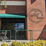 Brick building with a few tables, chairs and umbrellas in front of the wide, full-height windows. Several people are sitting at the tables. A railing and a hedge are visible in front of the landing. A circular logo on the bricks between two windows says The Crafted Scone.