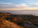 Looking down along a steeper hillside to a wide, shallow slobe with clusters of trees and dirt roads, and beyond that a coastline made of several coves and points. The hillside is lit up almost orange from the sunlight, and the trees (some darker green, some orange) case long shadows to the left. The ocean is calm, reflecting the pastel blues, yellows and pinks of a sky with feathery clouds, and a low island is silhouetted at the horizon.