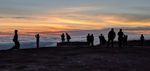 Several groups of people stand on shadowed, rocky ground, silhouetted against the sky...the top of a rolling cloud layer visible below them. Above, the sky is crisscrossed by clouds variously shadowed gray or lit yellow or orange, fading to pale blue higher up, but showing little clue to where the setting sun must be.