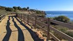 A wide, curving path between two wooden fences stretches into the distance. To the right, a dropoff leads to the ocean. Palms and other trees are visible in the distance on the land side. A lighthouse sits atop rugged cliffs in the distance, and beyond that, the silhouette of an island rises above the horizon.