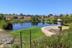 Small lake or large pond. At the near end is large pillar-type drain. A baseball field is visible at the far shore, next to some thicket of plants, unidentifiable from this distance. Beyond that, brown-green hills with houses on top and dirt trails winding along the hillside.
