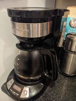 A simple drip coffee maker on a fake-granite countertop. The base is flared and has a handful of buttons on either side of a small LCD display. The coffee pot is round, glass, tapered at the top, with a black plastic handle and lid. It's filled up to about the 6-cup line. Above the pot there's a cylindrical holder for the coffee filter. Most of the case is black plastic, same as the handle and lid, with metal veneer wrapped around the filter basket and plated around the buttons on the base. There's a plastic gauge on the side, lines marked in Sharpie with two of them labeled 10 and 6. A cylindrical metal coffee grinder stands just behind it on the counter.