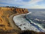 Red rugged cliffs rise above a pebbly beach while waves gently roll in from the ocean. A white column of a lighthouse sits atop the cliff, flanked by palm trees.
