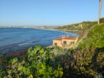 A squat, beige building with a tile roof has glommed onto the side of the bluffs rising from a cove. The shore curves away around a bright blue ocean, the scrub clinging to the bluffs turning the hillside a mottled green. A nearby plant with crinkly, fan-like leaves screens the rocky shore below. Past the building, toward the middle of the cove, the shallow water along the beach is smooth enough to reflect the steep hillside above. On the far side, the bluffs gradually lower until the shoreline barely shows a strip of green between the beach and the jumble of a coastal town. Off in the distance, a layer of haze separates the blue sky from the low silhouette of a mountain range.
