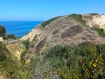 A steep, narrow channel curves around a hill beyond which you can see a blue triangle of ocean. The hill is brownish-yellow, but green plants grow in the foreground and down in the channel. Off to the left, if you look closely, you can see a stream at the bottom of the channel.