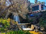 A waterfall about the height of a house, its lowest step clearly a flat block of concrete, drops from behind a sparsely-leaved tree and past a short building with enclosed balconies. Past the edges of the waterfall you can see a pair of spreading concrete walls with water stains and crawling vines. Grasses, mallows and iceplants grow around the edges and up to the foreground.