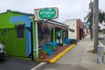 Small building painted green next to a sidewalk lined with palm trees. Dried palm fronds cover an awning, and two small round tables, each with a pair of chairs, flank an open glass door.
