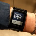 Close-up of a blocky digital watch on my wrist, showing the time (5:58) and date (October 26), with a banner across the lower third of the display indicating that sunset is in 9 minutes and an icon showing two clouds to indicate the weather.