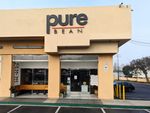 A corner storefront in a one-level flat building with parking spaces, a long bench under the porch in front of the full height windows, and a pair of glass doors. Above the entryway is the word 'pure' in very big lowercase letters, 'BEAN' below it in line with the descender on the 'p'.