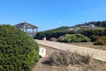 A stone path runs diagonally from one corner of the frame toward an octagonal gazebo, half-hidden behind a bright green bush. A hill rises in the distance below a bright blue sky.