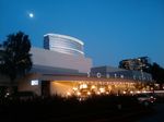 A wide, low building with an overhang lit up by *lots* of lights, against a dark blue sky. Two taller glass-walled office buildings are visible behind it, as is the bright splotch of the moon. Silhouettes of trees and a spiky hedge frame the sides and bottom of the frame. The words SOUTH COAST can be seen running along the overhang.