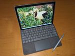 An ultra-thin tablet display with rounder corners, attached to a very flat, compact  keyboard. The keyboard is gray, with plastic keys and a trackpad surrounded by what looks like felt. A gray stylus with a small black nib rests diagonally across one of the empty corners next to the trackpad. The display is showing a Windows desktop with taskbar at the bottom and a wallpaper featuring a photo of plants and a pot in a garden.