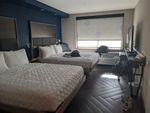 The hotel room. Two plain white queen-sized beds against a dark blue wall with brown headboards. There's just enough room to walk between the beds and a narrow shelf on which a flat-screen TV is perched. At the far end, light spills from a plain rectangular window, with a white shade mostly drawn down. Below it are a wall-unit air conditioner and a luggage stand. The floor is paneled in dark zig-zag planks that look like they might be wood, and a couple of bags and a pair of shoes are strewn about.