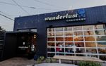 A boxy-looking storefont, possibly a former car repair shop cleaned up, with a large multi-paned glass window suspiciously shaped like a garage door. People are milling around inside, while a table stands empty in front of the building. A flat awning extends past a door, and a neon sign above the window proclaims Wanderlust Creamery with a simple ice cream cone.