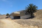 Flat area in front of a hillside with mostly dry grass. A wide concrete structure is built into the hill and flanked with palm trees. A dark opening is visible in the middle of the bunker, with a round concrete awning protecting it from above.