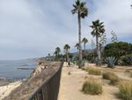 Looking along a railing outdoors on a sunny day. To the left, the ground drops off steeply. A beach and the ocean are visible below. To the right there are tufts of grass, palm trees, bushes and a paved path. A line of benches and plaque stands runs between the railing and the path.