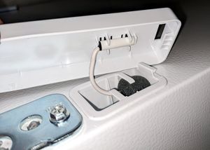 The plastic hinge cover has been lifted off of the metal hinge. A small cylinder is clipped into the cover, connected to a wire running into a tiny basket indented set into the top of the refrigerator.